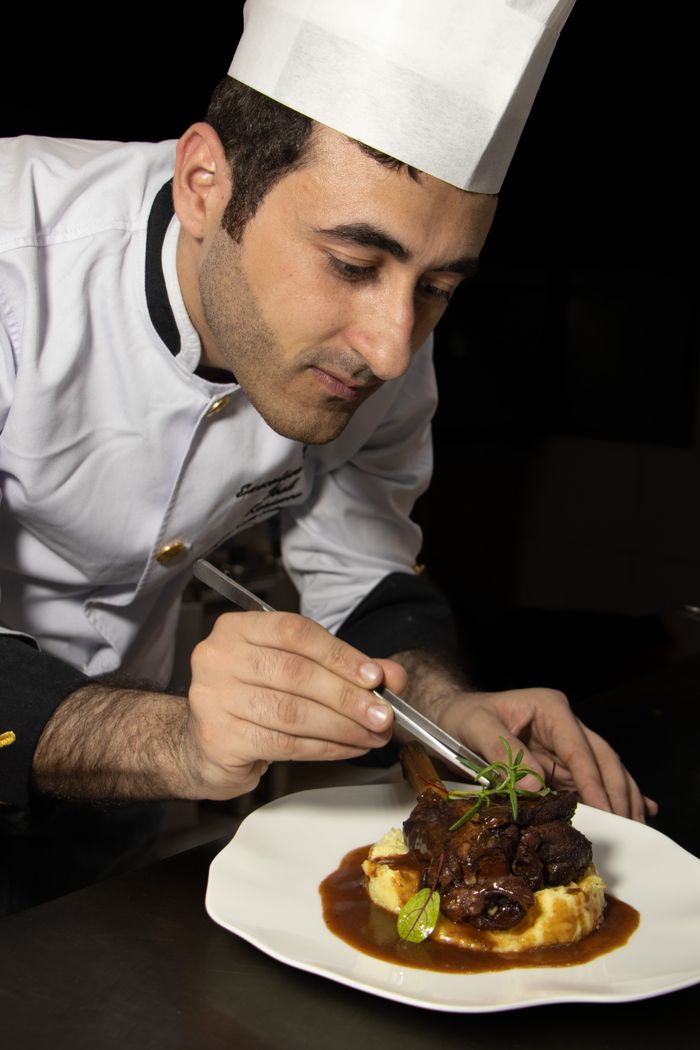 services-01 Mid adult chef skillfully garnishing meat dish with precision on a plate in studio setting.