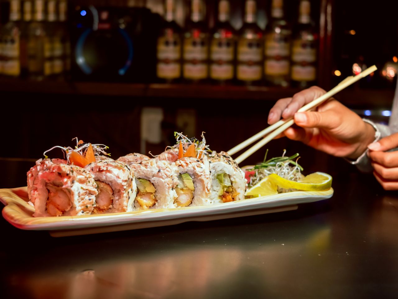 services-06 Close-up of gourmet sushi rolls with chopsticks in a cozy bar setting, highlighting fresh ingredients.